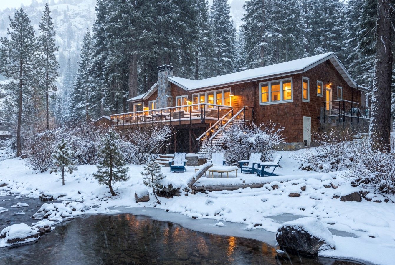 A cabin with a deck, a stone fireplace, and a pond in a snowy forest.