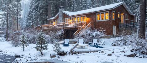 A cabin with a deck, a stone fireplace, and a pond in a snowy forest.