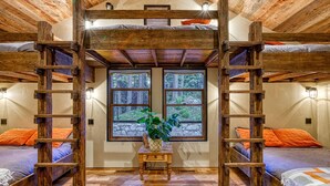 A rustic room with wooden beams, bunk beds, a central table with a plant, and large windows with a forest view.