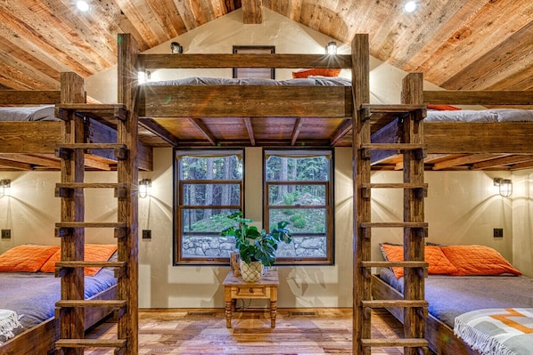 Una habitación rústica con vigas de madera, literas, una mesa central con una planta y grandes ventanales con vista al bosque.