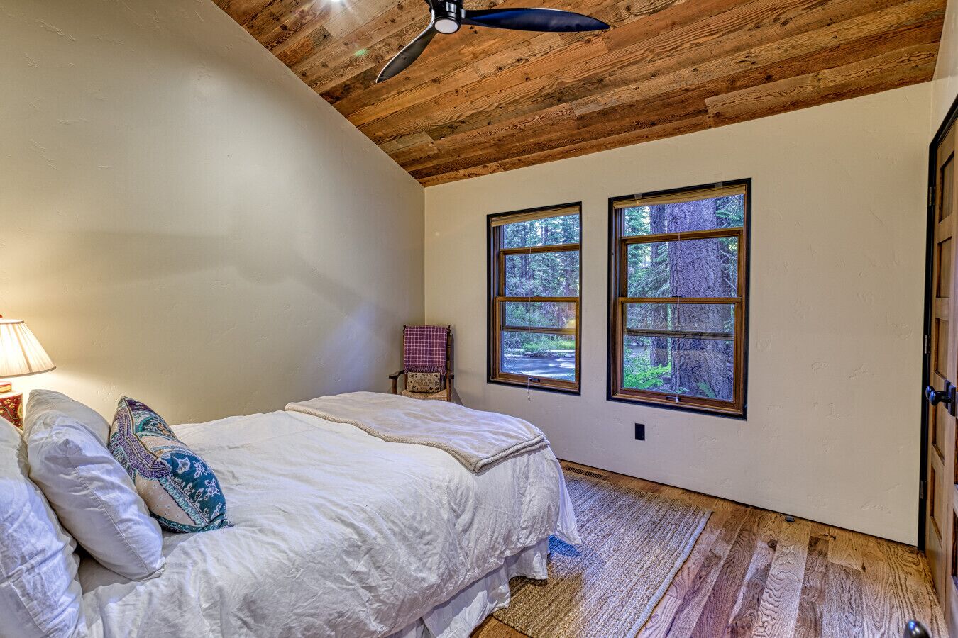 Un dormitorio con techo de madera, dos ventanas con vistas a un bosque, una cama con ropa blanca, una silla con funda a cuadros y una lámpara a la izquierda.