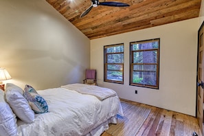 Un dormitorio con techo de madera, dos ventanas con vistas a un bosque, una cama con ropa blanca, una silla con funda a cuadros y una lámpara a la izquierda.