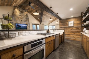 A modern kitchen with a stone accent wall, wooden beams, and a large island with appliances. - Truckee Riverfront Home | Minutes to Palisades (Tahoe City)