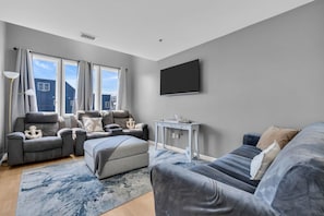 Een moderne woonkamer met een grijze bank, een grijze fauteuil, een klein tafeltje en een groot raam met gordijnen.