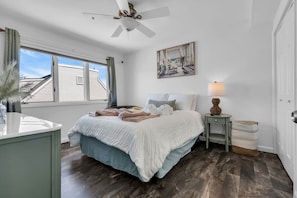 Une chambre à coucher avec un lit, un ventilateur de plafond, une fenêtre, un tableau, une table de chevet et une lampe.