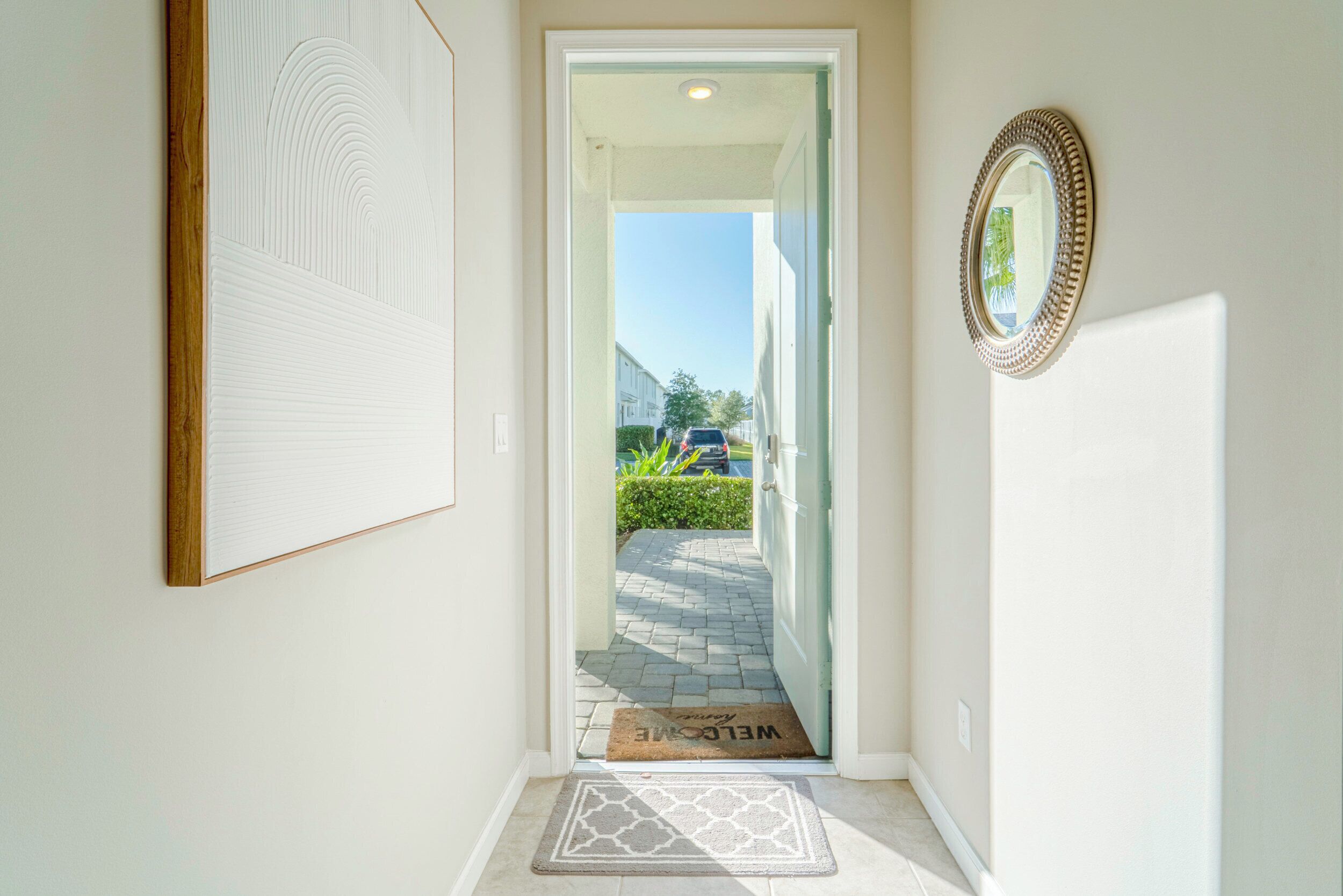 A clean, modern hallway with a welcome mat, a framed mirror, and a door leading to a residential street.