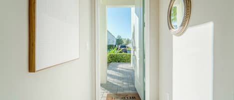 A clean, modern hallway with a welcome mat, a framed mirror, and a door leading to a residential street.