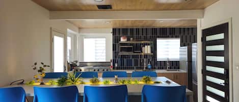 A modern kitchen with a gas stove, a sink, and a refrigerator. There is a dining area with blue chairs and a table set for four.