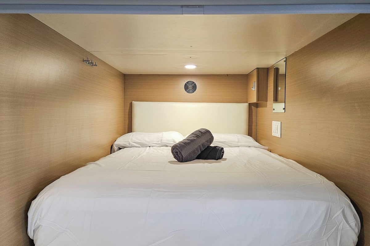 A single bed with white bedding, a rolled towel, a headboard, a wall-mounted light, and a mirror.