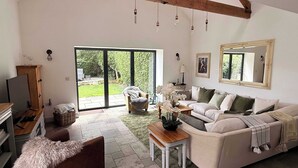 A spacious living room with a large sliding glass door leading to a garden, a sofa set, a wooden coffee table, and a television on a wooden stand.