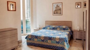 Une chambre à coucher comprenant un lit, une commode, une table de chevet, une lampe, un tableau représentant un bâtiment accroché au mur, et une porte donnant sur un balcon.