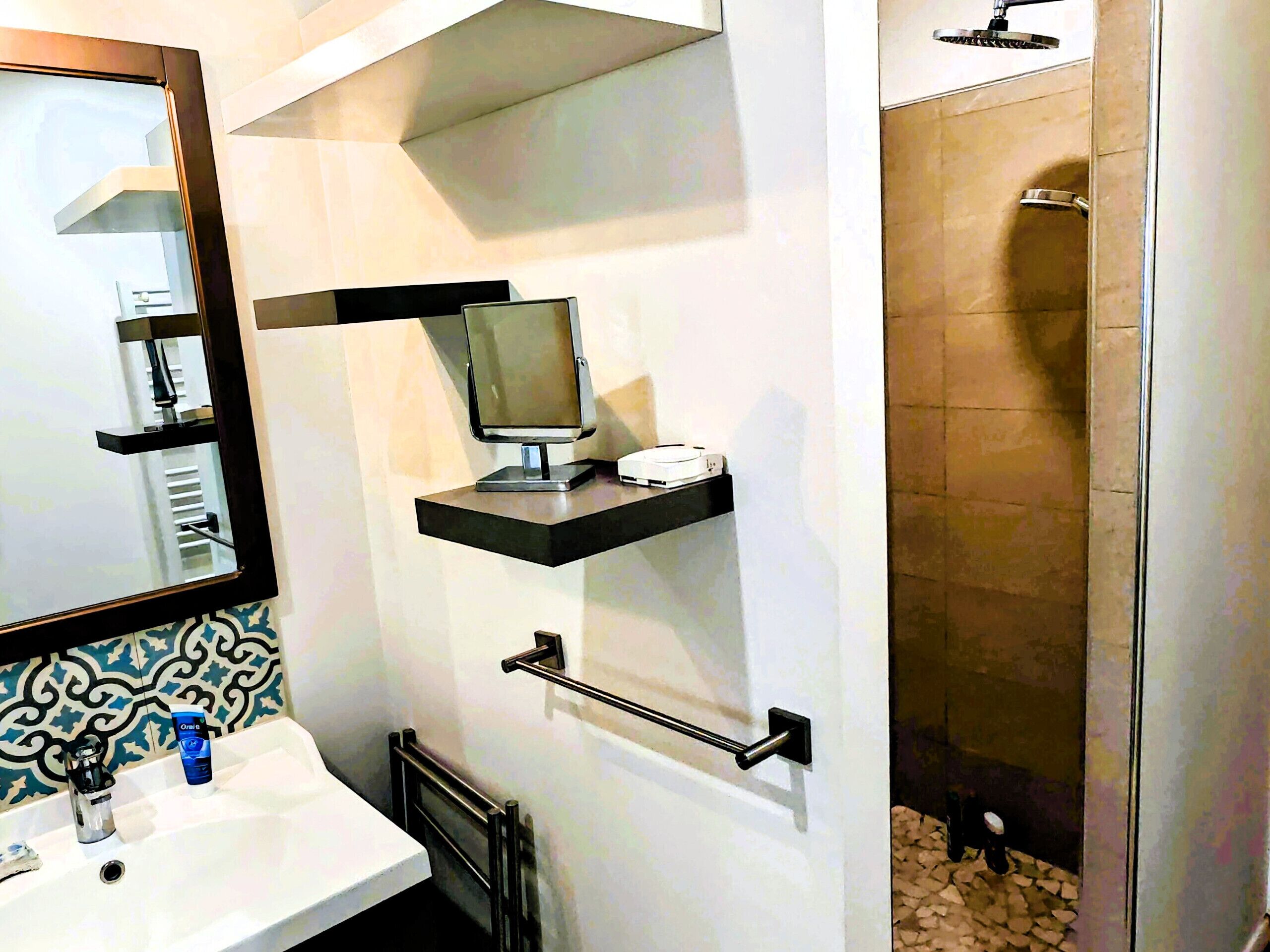 A bathroom with a white sink, a mirror, a toilet, a shower, and a small shelf with a computer and a cup.