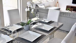 Ett modernt matområde med ett glasbord för fyra personer, vita stolar och en grå soffa i bakgrunden.