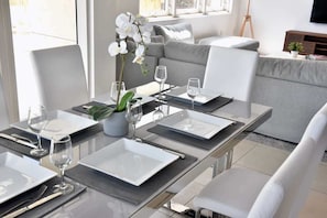 Ein moderner Essbereich mit einem Glastisch, gedeckt für vier Personen, weißen Stühlen und einem grauen Sofa im Hintergrund.