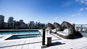 Una piscina en la azotea con un horizonte urbano de fondo, una zona de estar junto a la piscina y un cielo despejado.