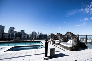 Ein Pool auf dem Dach mit Stadtblick im Hintergrund, ein Sitzbereich am Pool und ein klarer Himmel.