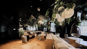 Un área de descanso moderna con suelo de madera, un sofá grande, mesas redondas, plantas colgantes y vistas a la ciudad.