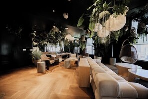 Eine moderne Lounge mit Holzböden, einer großen Couch, runden Tischen, hängenden Pflanzen und Blick auf die Stadt.