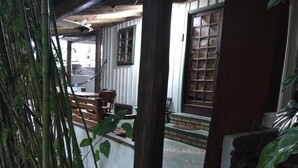 Un espacio al aire libre cubierto con un banco de madera, una mesa y una silla. Se observan ventanas con marcos de madera y una puerta.