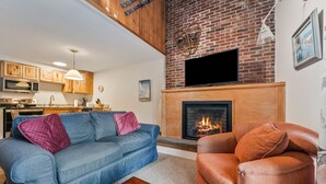 A living room with a fireplace, a blue sofa, a brown armchair, and a kitchen in the background. - Walk to Attitash in 2 Mins | Cozy Mountain Condo (Bartlett)