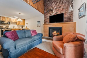 A living room with a fireplace, a blue sofa, a brown armchair, and a kitchen in the background. - Walk to Attitash in 2 Mins | Cozy Mountain Condo (Bartlett)