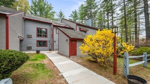 A two-story building with a red door and windows, a concrete pathway, and a yellow flowering bush. - Walk to Attitash in 2 Mins | Cozy Mountain Condo (Bartlett)