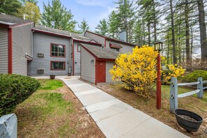 A two-story building with a red door and windows, a concrete pathway, and a yellow flowering bush. - Walk to Attitash in 2 Mins | Cozy Mountain Condo (Bartlett)