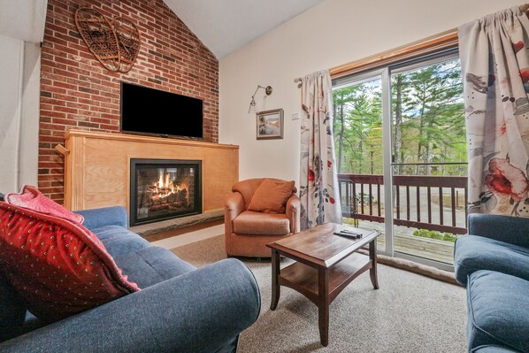A living room with a brick fireplace, a television mounted above it, a sofa, a chair, and a balcony with a view of trees. - Walk to Attitash in 2 Mins | Cozy Mountain Condo (Bartlett)