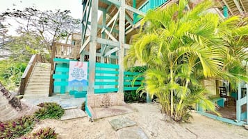A beachside villa with a staircase leading to a wooden deck, surrounded by tropical plants and a sign indicating a beachside location.