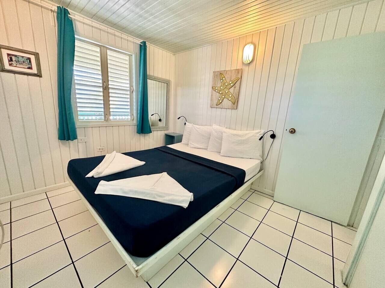 En enkeltseng med hvide sengetøj, et turkis gardin, en stjernefisk som dekoration på væggen og et hvidt skab.