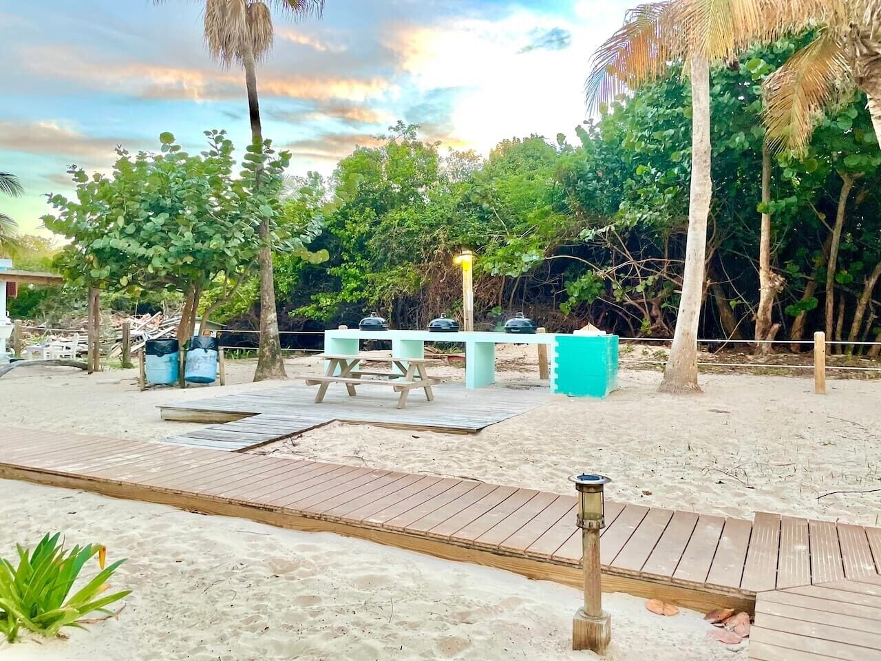Ein Strandbereich mit Sand, einer Holzterrasse, Picknicktischen und einem Grill.