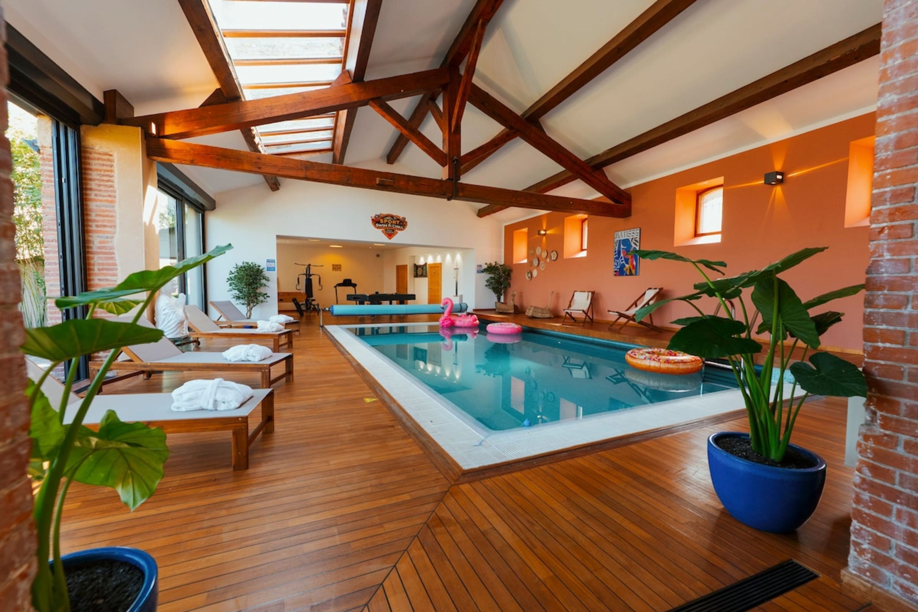 A spacious indoor swimming pool with wooden flooring, lounge chairs, and potted plants.