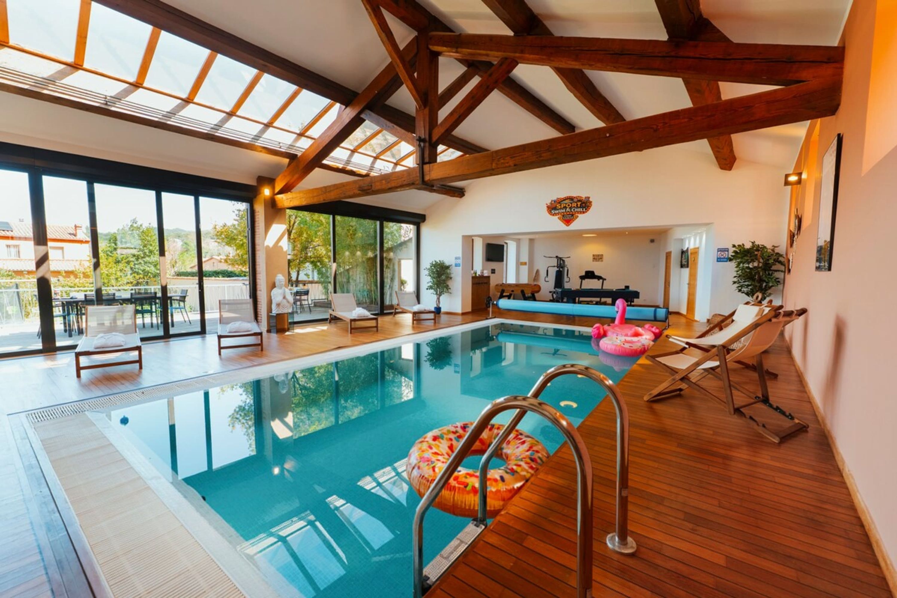 An indoor swimming pool with wooden beams, lounge chairs, and a view of the outside.