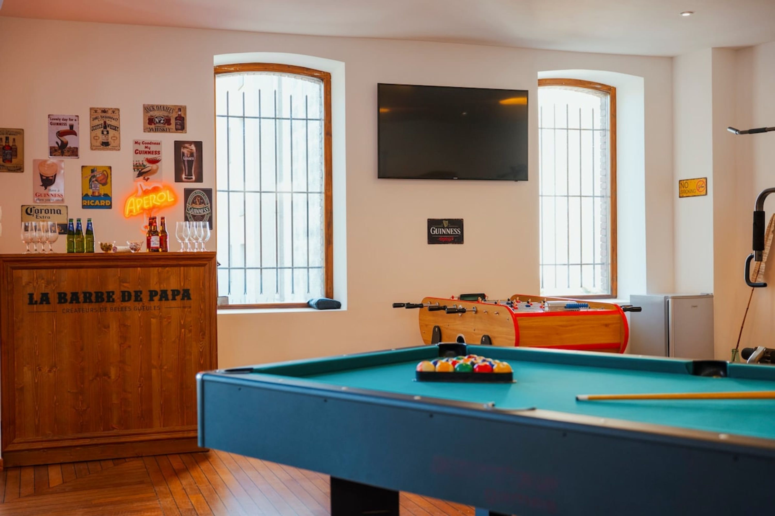 A pool table with a neon sign, a foosball table, and a bar area with a sign reading 'LA BARBE DE PAPA'.