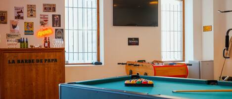 A pool table with a neon sign, a foosball table, and a bar area with a sign reading 'LA BARBE DE PAPA'.