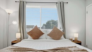 A bed with a white quilt, two brown pillows, a beige blanket, a window with blinds, and two bedside lamps on wooden nightstands. - Townie at the Foothills | Hop, Skip & Hike (Fort Collins)