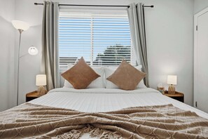 A bed with a white quilt, two brown pillows, a beige blanket, a window with blinds, and two bedside lamps on wooden nightstands. - Townie at the Foothills | Hop, Skip & Hike (Fort Collins)