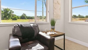 Ein Zimmer mit Ledersofa, einem kleinen Tisch mit Buch und Topfpflanze sowie großen Fenstern mit Blick auf eine grüne Landschaft.