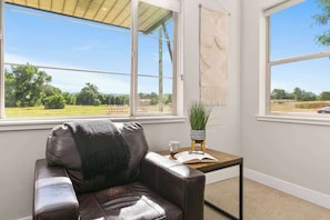 A room with a leather sofa, a small table with a book and a potted plant, and large windows offering a view of a green landscape. - Rocky Retreat Near Old Town | Hot Tub | Fireplace (Fort Collins)