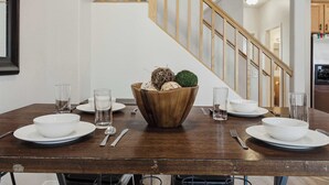 Una mesa de comedor dispuesta con cuencos, platos y cubiertos blancos, un centro de mesa de madera y una escalera al fondo.