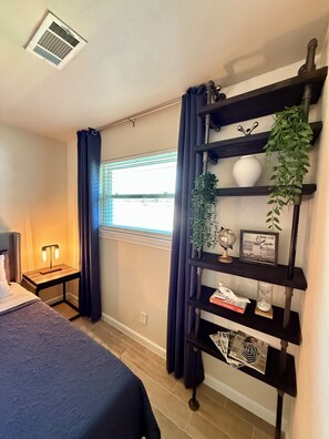 A bedroom with a bed, a nightstand, a shelf with decorative items, a window with curtains, and a ceiling vent. - Cozy & Charming Stay Min from Hobby, NRG, Downtown, NASA (Houston)