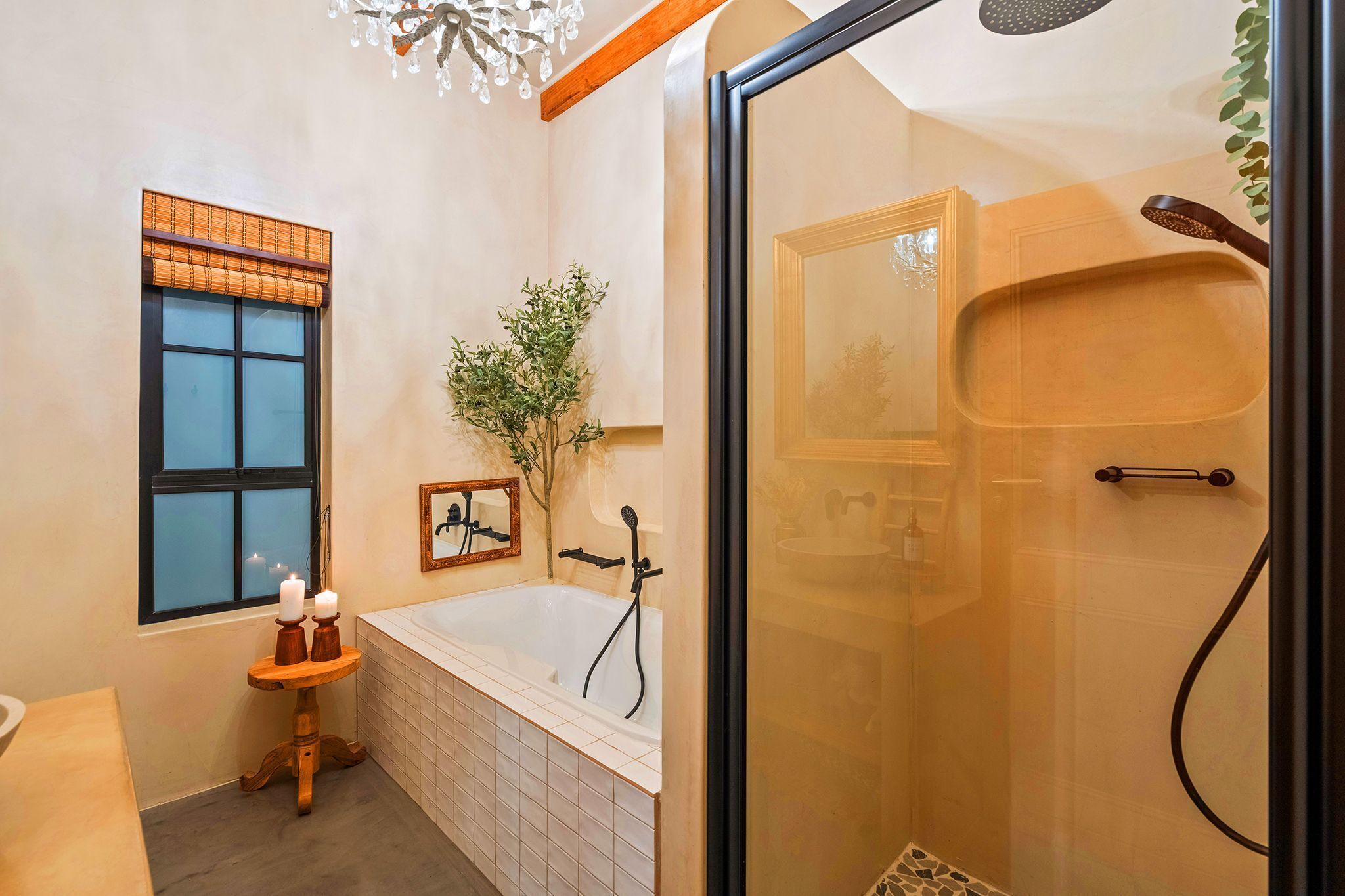 Une salle de bain avec une baignoire, une douche en verre, une fenêtre avec un store en bambou, une petite table avec des bougies et une plante en pot.