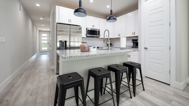 A modern kitchen with white cabinets, a stainless steel refrigerator, a microwave, and a sink. There are three black bar stools and pendant lights above the counter. - 4BR Pet-Friendly Beach Home | Walk to Beach & Jacksonville Beach Pier | Fenced Yard (Jacksonville Beach)