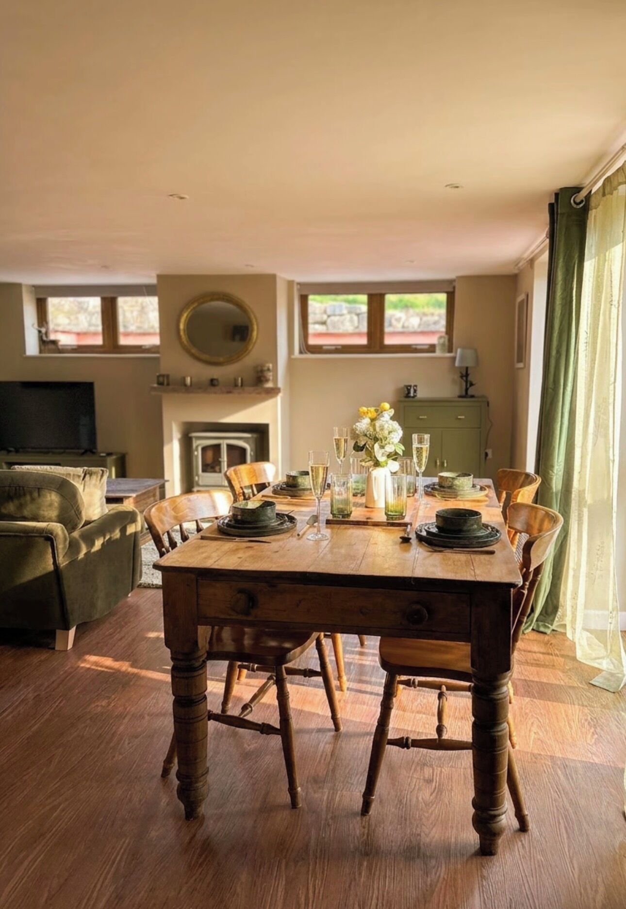 Een eethoek met een houten tafel voor vier personen, omringd door stoelen. Er is een open haard met een schoorsteenmantel, een televisie links en een raam met gordijnen rechts.