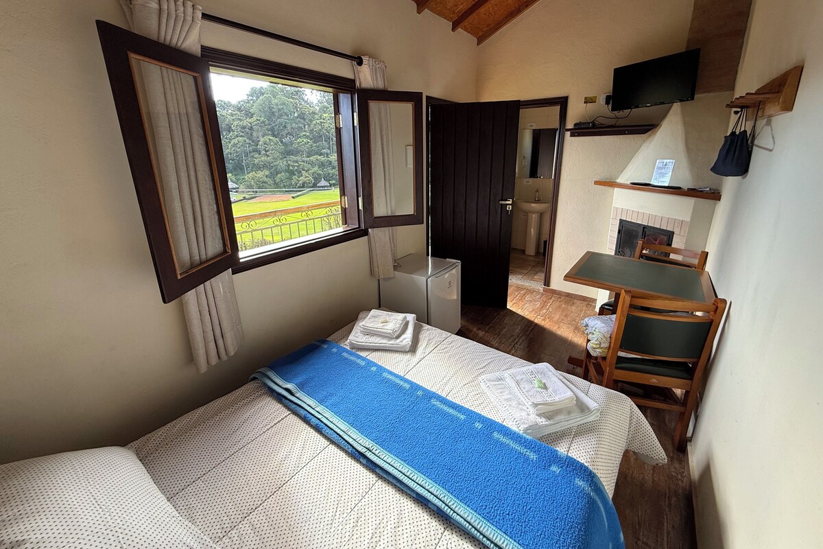 Una camera da letto con un letto, una finestra con tende, una scrivania, una sedia e un bagno visibile attraverso una porta aperta.
