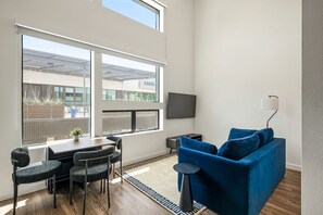 A modern living room with a dining area, large windows, a flat-screen TV, and a blue sofa. - Vibrant DT ATX Loft | Near Rainey St (Austin)