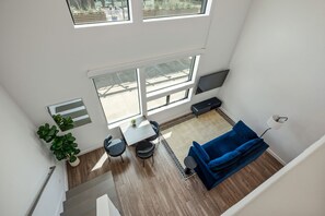 A high-ceilinged room with a large window, a flat-screen TV, a blue sofa, a small table, and potted plants. - Vibrant DT ATX Loft | Near Rainey St (Austin)