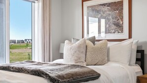 A bedroom with a large bed, a view of a grassy field, and a framed picture on the wall. - Mallard Cove | Pond Views | Pets Ok | Playground (Calgary)