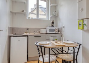 Een compacte keuken met een koelkast, magnetron en koffiezetapparaat. Er is een eettafeltje met twee stoelen.