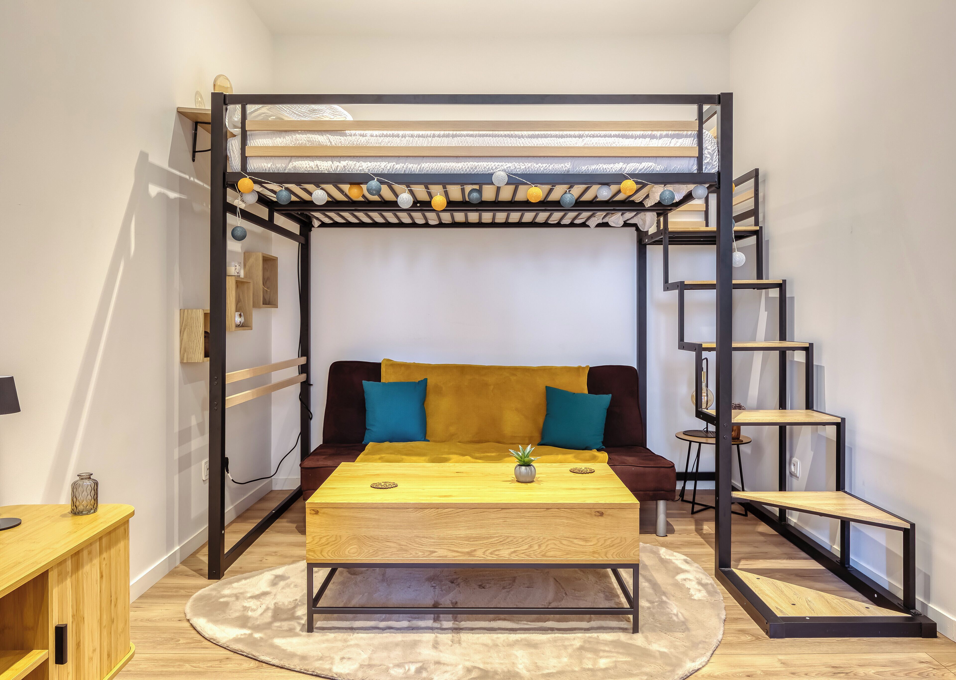 Una habitación con literas, un sofá cama, una mesa de centro de madera, una planta pequeña, una alfombra redonda y una escalera.